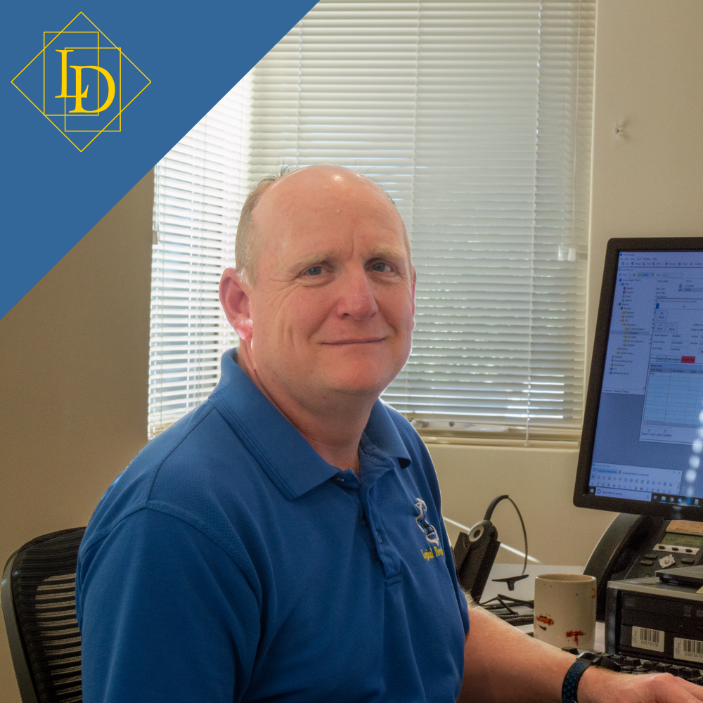 It's a photo of Graeme. He's sitting at his desk and is slightly looking from left to the right. He faces the camera with a neutral face. Behind him are some of the other workstations. In the top left corner is the Logical Developments logo in gold on blue. The Logical Developments logo which is a diamond with the letters L and D inside of it. The letters are offset from each other vertically while also overlapping horizontally. The each sit inside of their own rectangle.